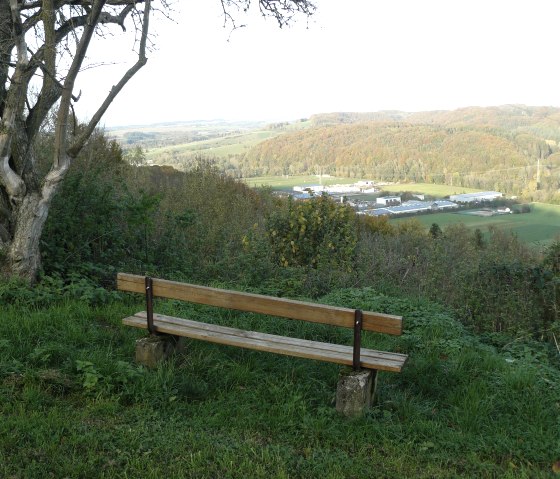 Wooden bench on a meadow with a view of a hilly landscape and an industrial area in the distance. There is a tree on the left of the picture., © Felsenland Südeifel Tourismus GmbH, Christian Calonec-Rauchfuss Wooden bench on a meadow with a view of a hilly landscape and an industrial area in the distance. There is a tree on the left of the picture., © Felsenland Südeifel Tourismus GmbH, Christian Calonec-Rauchfuss
