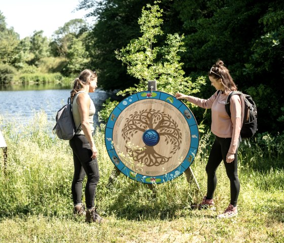 Two women with rucksacks look at a decorative wooden wheel on the shore of a lake, surrounded by green nature., © Eifel Tourismus GmbH, Dominik Ketz Two women with rucksacks look at a decorative wooden wheel on the shore of a lake, surrounded by green nature., © Eifel Tourismus GmbH, Dominik Ketz