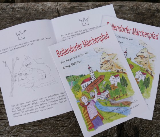 Coloring book pages of the Bollendorf fairytale trail with colorful illustrations and text about King Bollybur on a wooden table., © Felsenland Südeifel Tourismus GmbH Coloring book pages of the Bollendorf fairytale trail with colorful illustrations and text about King Bollybur on a wooden table., © Felsenland Südeifel Tourismus GmbH