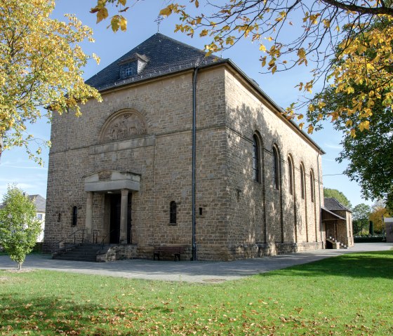 The church of St. Hubertus in Wolsfeld, surrounded by autumnal trees, stands in a green park. The building is made of stone with a gabled roof., © TI Bitburger Land The church of St. Hubertus in Wolsfeld, surrounded by autumnal trees, stands in a green park. The building is made of stone with a gabled roof., © TI Bitburger Land