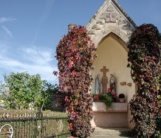Small chapel with statues, surrounded by autumn ivy. A wrought-iron gate and blue sky in the background., © TI Bitburger Land Small chapel with statues, surrounded by autumn ivy. A wrought-iron gate and blue sky in the background., © TI Bitburger Land