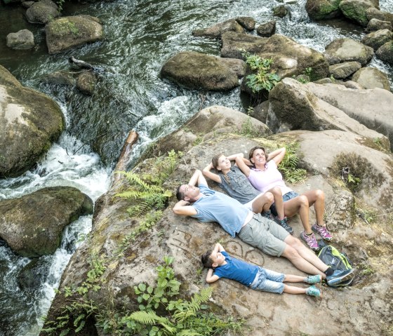 Eine Familie liegt entspannt auf einem großen Felsen inmitten eines fließenden Baches mit Wasserfällen und umgeben von üppigem Grün., © Felsenland Südeifel Tourismus GmbH Eine Familie liegt entspannt auf einem großen Felsen inmitten eines fließenden Baches mit Wasserfällen und umgeben von üppigem Grün., © Felsenland Südeifel Tourismus GmbH