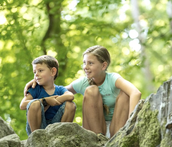 Zwei Kinder sitzen auf Felsen im Wald. Sie lächeln und schauen in die Ferne, umgeben von grünem Laub und natürlichem Licht., © Felsenland Südeifel Tourismus GmbH Zwei Kinder sitzen auf Felsen im Wald. Sie lächeln und schauen in die Ferne, umgeben von grünem Laub und natürlichem Licht., © Felsenland Südeifel Tourismus GmbH