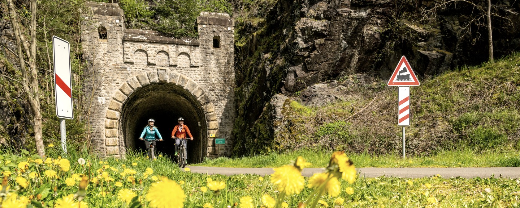 Zwei Radfahrer auf einem Weg vor einem alten Bahntunnel, umgeben von blühendem Löwenzahn und Verkehrsschildern., © Eifel Tourismus GmbH, Dominik Ketz Zwei Radfahrer auf einem Weg vor einem alten Bahntunnel, umgeben von blühendem Löwenzahn und Verkehrsschildern., © Eifel Tourismus GmbH, Dominik Ketz