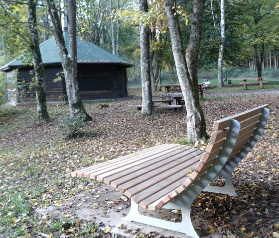 Wooden lounger and picnic tables in an autumnal forest with a hut in the background. Leaves cover the ground., © Felsenland Südeifel Tourismus GmbH, Christian Calonec-Rauchfuss Wooden lounger and picnic tables in an autumnal forest with a hut in the background. Leaves cover the ground., © Felsenland Südeifel Tourismus GmbH, Christian Calonec-Rauchfuss