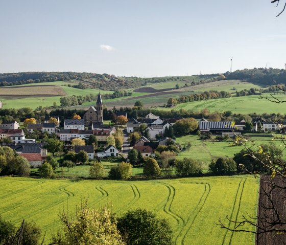 Panoramic view of the village of Alsdorf with a church in the center, surrounded by green fields and hills under a blue sky., © Tourist-Info Bitburger Land Panoramic view of the village of Alsdorf with a church in the center, surrounded by green fields and hills under a blue sky., © Tourist-Info Bitburger Land