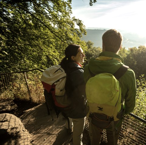Two hikers with rucksacks stand at a railing and look out over the sunny, wooded landscape., © Dominik Ketz Two hikers with rucksacks stand at a railing and look out over the sunny, wooded landscape., © Dominik Ketz
