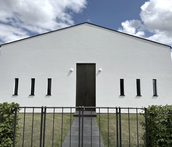 Modern white building with narrow windows and large wooden gate, surrounded by lawn and fence, under a blue sky with clouds., © Felsenland Südeifel, Anna Carina Krebs Modern white building with narrow windows and large wooden gate, surrounded by lawn and fence, under a blue sky with clouds., © Felsenland Südeifel, Anna Carina Krebs