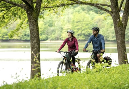 Two smiling cyclists ride along a path by the Bitburg reservoir, surrounded by green nature and trees., © Eifel Tourismus GmbH, Dominik Ketz Two smiling cyclists ride along a path by the Bitburg reservoir, surrounded by green nature and trees., © Eifel Tourismus GmbH, Dominik Ketz