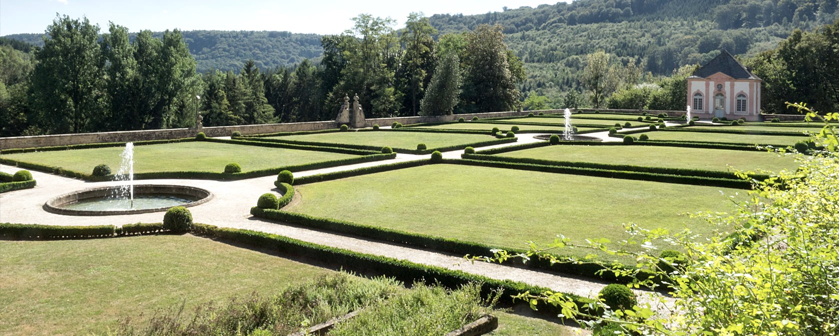 Ein gepflegter französischer Garten mit symmetrischen Hecken, Springbrunnen und einem Pavillon im Hintergrund, umgeben von Wald., © V. Teuschler Ein gepflegter französischer Garten mit symmetrischen Hecken, Springbrunnen und einem Pavillon im Hintergrund, umgeben von Wald., © V. Teuschler