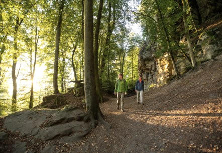 Two hikers are walking on a forest path along a rock face. The sun shines through the trees and bathes the scene in warm light., © Dominik Ketz Two hikers are walking on a forest path along a rock face. The sun shines through the trees and bathes the scene in warm light., © Dominik Ketz