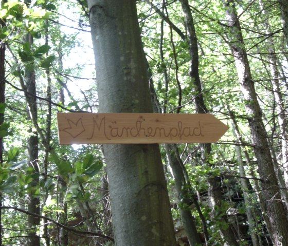 A wooden sign with the words 'Fairytale Trail' hangs on a tree in the forest and points the way., © Annette & Paul-Theo Colljung A wooden sign with the words 'Fairytale Trail' hangs on a tree in the forest and points the way., © Annette & Paul-Theo Colljung