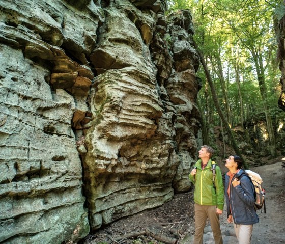Two hikers stand in front of impressive rock formations in a forest. The rocks have striking, layered structures. Sunlight filters through the trees. Two hikers stand in front of impressive rock formations in a forest. The rocks have striking, layered structures. Sunlight filters through the trees.