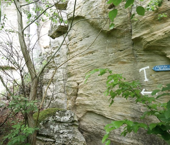 A rock face in the forest with a blue sign pointing the way to the Devil's Hole. Green leaves and trees surround the scene., &copy; Elke Wagner, Felsenland S&uuml;deifel Tourismus GmbH