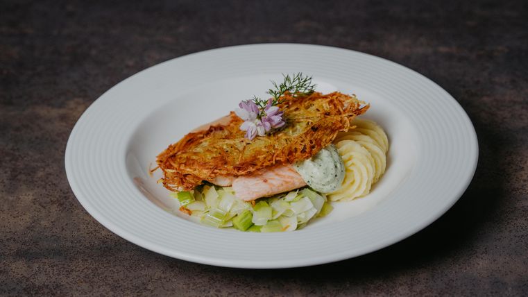 Ein schöner Teller mit gebratenem Lachs, umgeben von Püree und Lauch. Das Gericht ist mit Kräutern und einer essbaren Blume dekoriert.