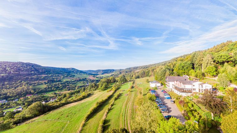 Die malerische Landschaft mit sanften Hügeln und üppigem Grün. Im Vordergrund steht das Waldhotel Sonnenberg, umgeben von Bäumen und einem Parkplatz.