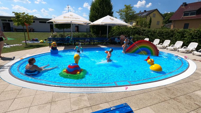 Ein schöner Pool mit bunten Schwimmspielzeugen und einem Regenbogenrutsche. Kinder spielen und relaxen im Wasser unter sonnigem Himmel.