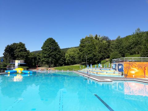 Ein Schwimmbecken an einem sonnigen Tag, umgeben von Bäumen. Im Hintergrund sind Liegestühle und ein Sprungturm zu sehen.