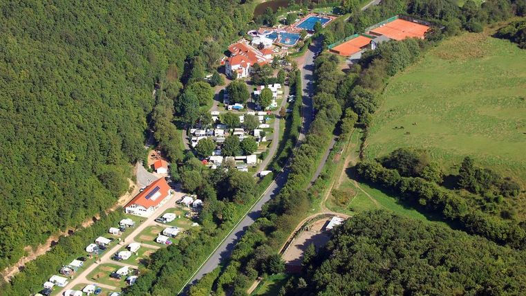 Eine Luftaufnahme eines Campingplatzes umgeben von Wald. Es gibt Wohnwagen, ein Schwimmbad und Tennisplätze in der Nähe.