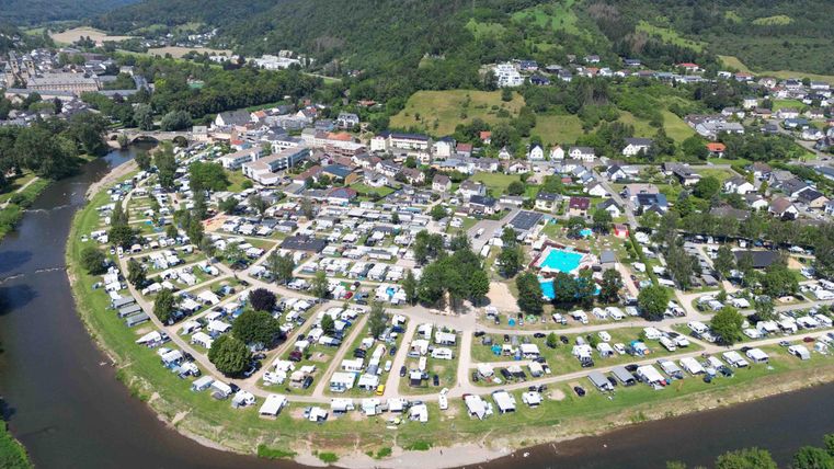 Eine Luftaufnahme eines Campingplatzes entlang eines Flusses. Umgeben von grünen Hügeln und Häusern ist der Platz mit Bäumen und einem Pool versehen.