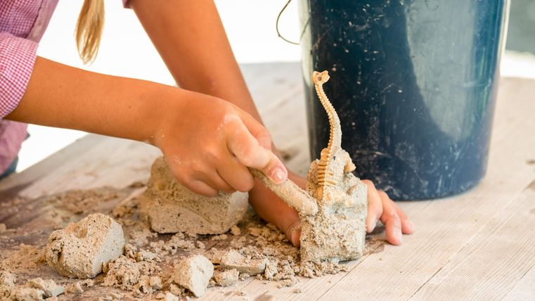 Ein Kind formt Sand mit einer Hand, während es eine kleine Figur hält. Im Hintergrund steht ein blauer Eimer.