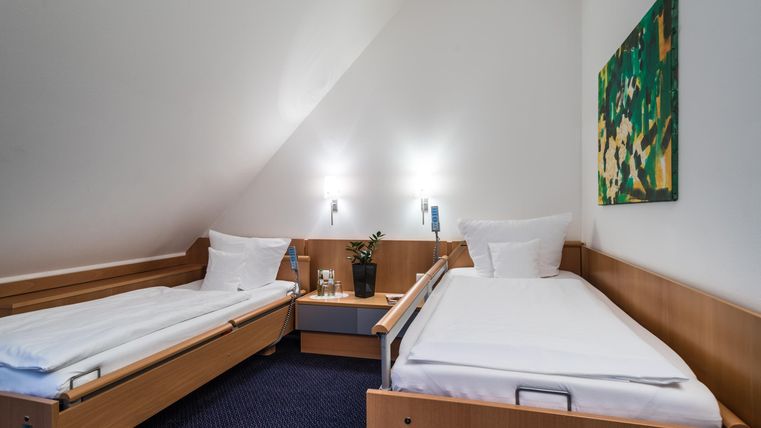 Ein einfaches Zimmer mit zwei Einzelbetten und modernen Möbeln. An der Wand hängt ein farbenfrohes Bild, und es gibt eine kleine Pflanze auf dem Nachtisch.