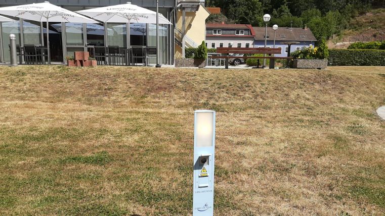 Ein heller, moderner Beleuchtungskörper steht auf einer Wiese vor einem Gebäude. Im Hintergrund sind Tischdecken und Sonnenschirme eines Außenbereichs sichtbar.