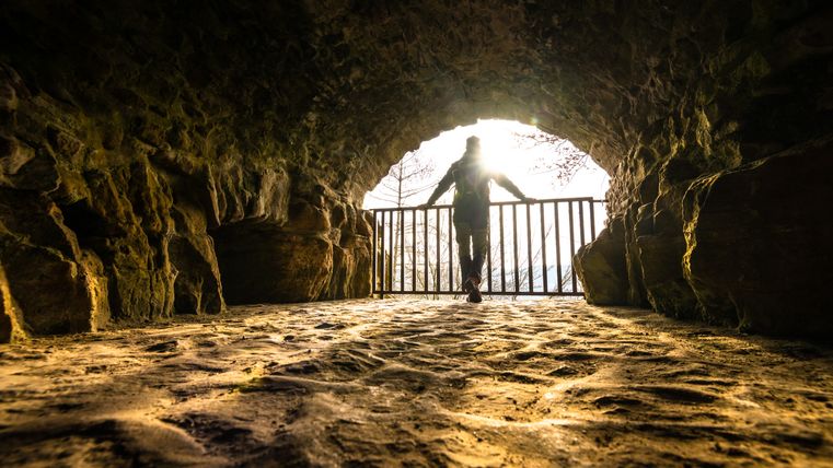Person steht in einem steinernen Tunnel, beleuchtet von goldenem Licht, mit Blick auf ein Gittertor.
