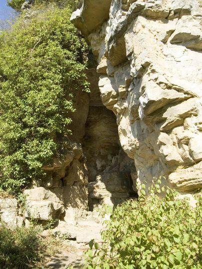 Eine natürliche Felsformation mit üppigem Grün und Schatten. Der Eingang zu einer Höhle ist sichtbar.