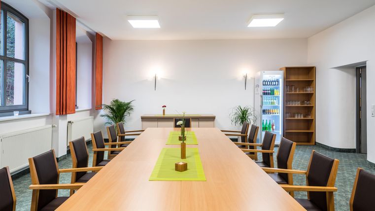 Ein moderner Besprechungsraum mit einem langen Tisch und Stühlen. Die Wände sind hell und es gibt Pflanzen sowie Bücherregale im Hintergrund.