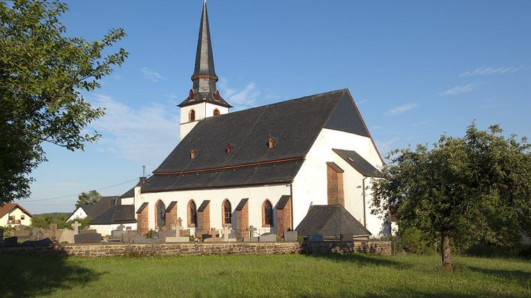 Eine alte Kirche mit einem hohen, spitzen Turm in einer ruhigen ländlichen Umgebung. Umgeben von Wiesen und Bäumen, strahlt sie eine friedliche Atmosphäre aus.