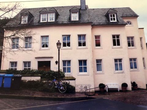 Ein mehrstöckiges, helles Gebäude mit vielen Fenstern. Vor dem Gebäude stehen ein Fahrrad und eine Bank, umgeben von einigen Pflanzen.