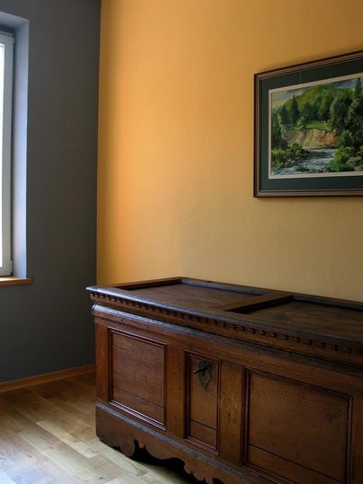 Ein gemütlicher Raum mit einer großen Holztruhe und einem Gemälde an der Wand. Die Wände sind in sanften Farben gestrichen, und das Licht fällt durch das Fenster.