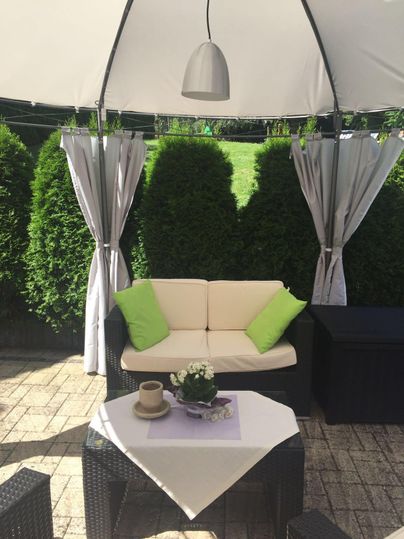 Ein gemütlicher Gartenbereich mit einem Sofa und Kissen in Grün. Ein Tisch mit einer Pflanze und einem Kaffeebecher sorgt für eine einladende Atmosphäre.