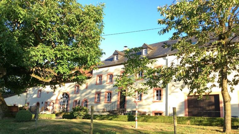 Ein großes, helles Gebäude mit roten Fenstern und einem schönen Garten. Rund um das Haus wachsen grüne Bäume und der Himmel ist klar und blau.