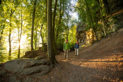Zwei Personen wandern auf einem Waldweg nahe einer Felswand bei Sonnenuntergang.
