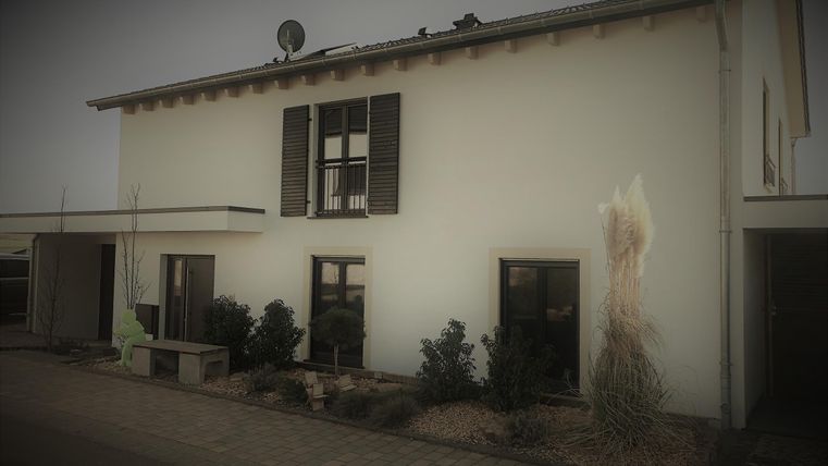 Een modern huis met witte muren en donkere luiken. Er zijn planten aan de voorkant en een schotelantenne op het dak.