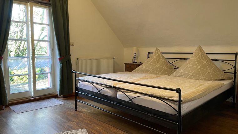 Ein gemütliches Schlafzimmer mit einem großen Bett und zwei Kissen. Fenster mit Vorhängen lassen Tageslicht herein.