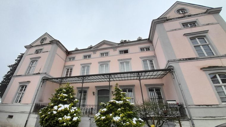 Ein großes, historisches Gebäude in zartem Pink mit grauen Akzenten. Vor dem Gebäude befinden sich schneebedeckte Sträucher.