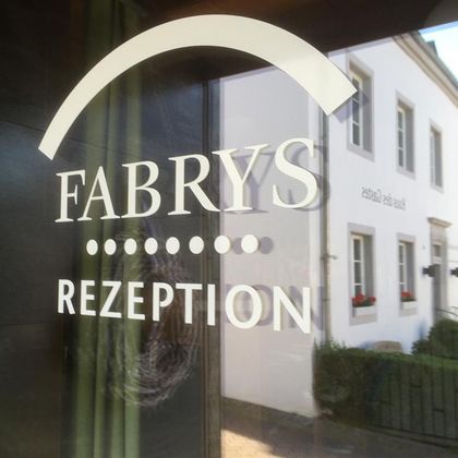 Ein Fenster mit dem Schriftzug „FABRYS REZEPTION“. Im Hintergrund ist ein Gebäude mit Blumenfenstern zu sehen.