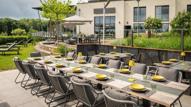 Ein eleganter Außenbereich mit einem langen Tisch, der für ein Essen gedeckt ist. Im Hintergrund ist ein modernes Gebäude und ein gepflegter Garten zu sehen.