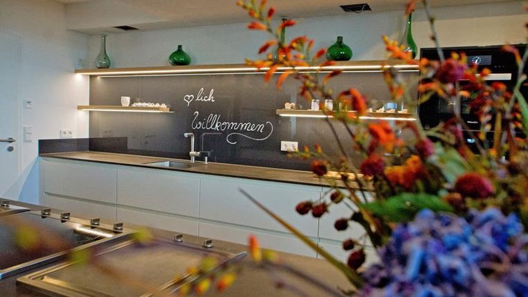 Eine moderne Küche mit eleganten Regalen und einer Tafel, die "Willkommen" schreibt. Im Vordergrund sind bunte Blumenarrangements sichtbar.