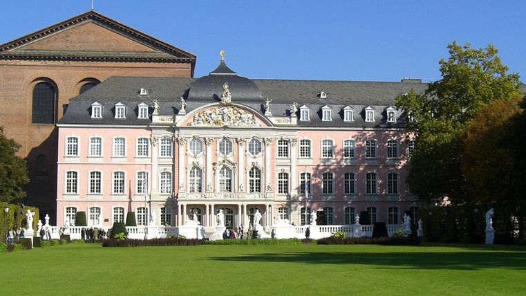 Ein majestätisches Schloss mit rosa Fassaden und kunstvollen Verzierungen. Die weitläufige Grünfläche und der klare Himmel schaffen eine einladende Atmosphäre.