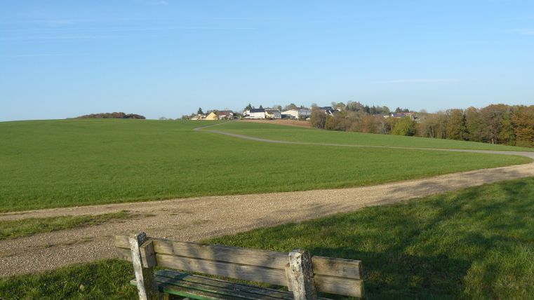 Blick auf eine grüne Wiese mit einem Weg und einer Bank im Vordergrund, im Hintergrund ein Dorf unter blauem Himmel.