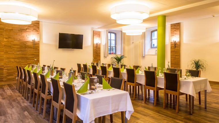 Ein stilvoll eingerichteter Veranstaltungsraum mit langen Tischen und Stühlen. Die Tische sind mit weißen Tischtüchern und grünen Servietten dekoriert.