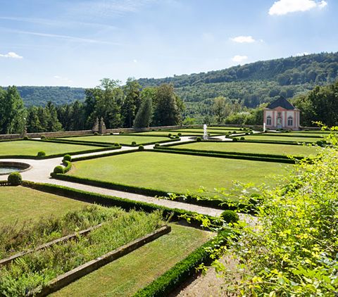 Französischer Garten mit geometrischen Hecken und Rasenflächen, im Hintergrund ein kleines Gebäude und bewaldete Hügel.