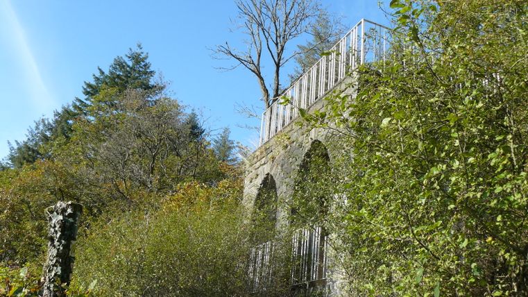 Steinmauer mit Geländer in bewaldeter Umgebung unter blauem Himmel.