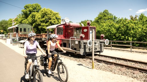 Zwei Radfahrerinnen auf einem Radweg neben einer alten Lokomotive im Eisenbahnmuseum Pronsfeld, umgeben von grünen Bäumen., © Eifel Tourismus GmbH, Dominik Ketz