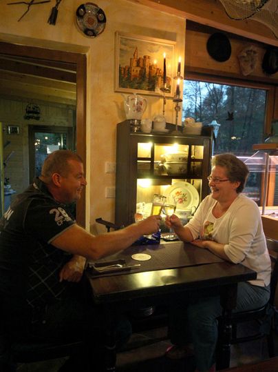 Ein Mann und eine Frau sitzen an einem Tisch in einem gemütlichen Raum. Sie scheinen sich zu unterhalten und lachen, während im Hintergrund dekorative Gegenstände zu sehen sind.
