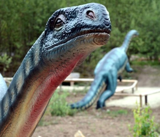 Ein Dinosauriermodell im Dinopark, umgeben von grüner Vegetation. Im Hintergrund ist ein weiteres Dinosauriermodell zu sehen.